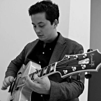 Lucian Gray is standing, wearing a blazer while playing guitar in a black and white photo.