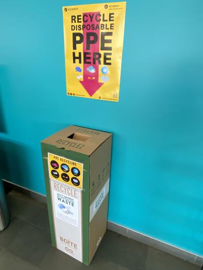 A sign with a red arrow pointing downward that reads recycle disposable PPE here. Underneath the sign is a box with a sign that 
