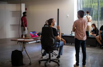 A person sits at a desk with a laptop computer while another person talks to him.