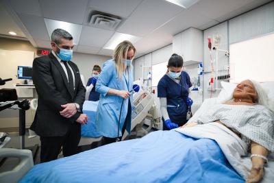 Ross Romano and a Nursing instructor watch as a student in blue scrubs tends to a mannequin in a simulation