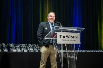 Ray Chateau speaks into a microphone while standing in front of a number of award statues.