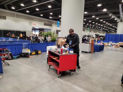 A person reaches for items on a workbench filled with tools and equipment as other people work in the background.