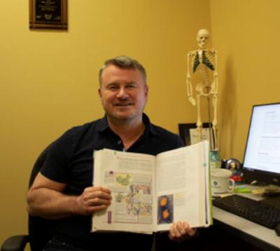 Professor Ron Stewart sits at his desk holding open a science textbook with a skeleton model in the background