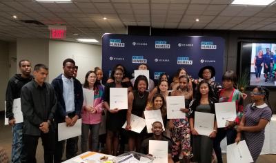 A group of 20 people hold up pieces of paper with writing on them as they stand in front of a blue sign that reads We Are Humber