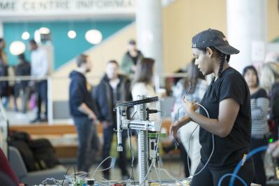 A person wearing a backwards ball cap and braided pigtails manipulates a long wire at a table full of mechatronics equipment.