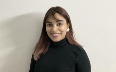 Anshika Na smiles in a headshot against a muted white wall, wearing a black turtleneck. Her hair brown hair is shoulder-length