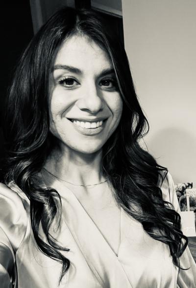 Atash Khosrorad smiles in a selfie headshot, wearing a satiny blouse and her long brown hair in loose curls. 