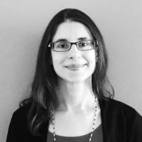 Natalie Stevens smiles in front of a grey background. She is wearing glasses and a dark cardigan. The photo is black and white