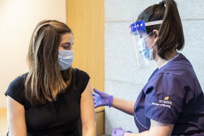 A Humber Nursing student wearing a face shield prepares to vaccinate a person with long brown hair. They are both wearing masks
