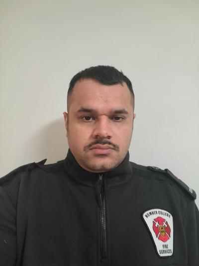 Munir Raza is wearing a black jacket with a firefighter's patch on the left breast. He is straight-faced and has a dark moustach