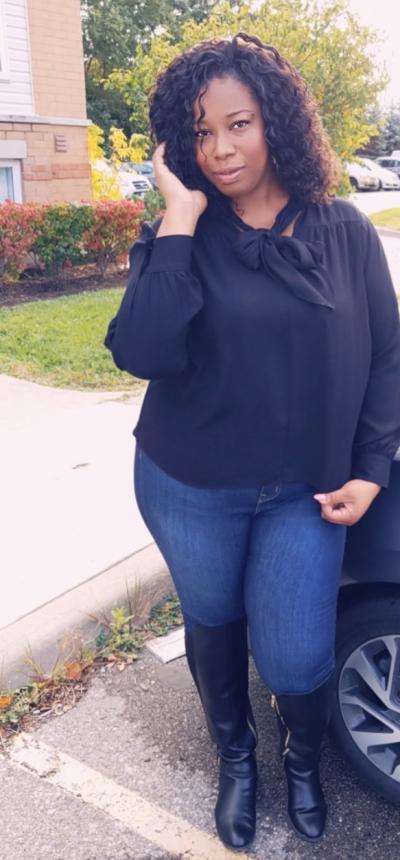 Meshanda Ellison poses in front of a car wearing jeans and a black shirt. Her hand is touching her curls softly