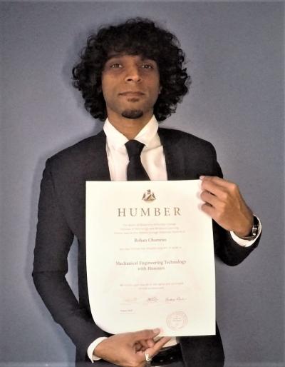 Rohan Chamroo is standing in front of a grey background wearing a suit, smiling and holding his credential