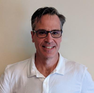 Professor Culum Canally smiles in a headshot, wearing a white button-up and black glasses in front of a beige wall.