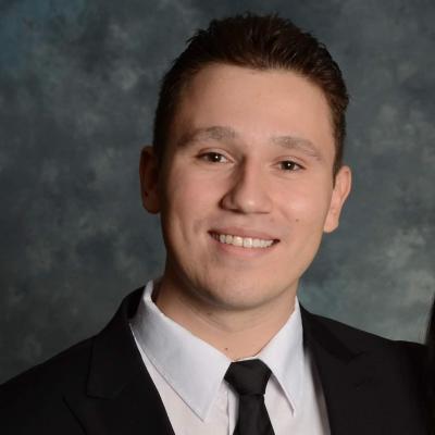 Paul-Michael Broz is smiling in front of a grey background, wearing a white shirt and black jacket and tie