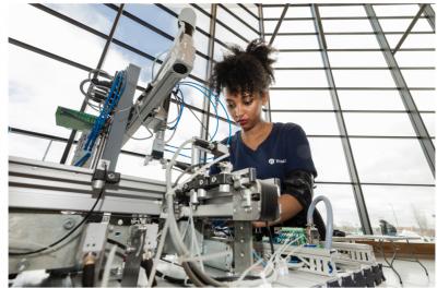 A woman looks down at the manufacturing machine she is using. She is backlit by a wall of windows