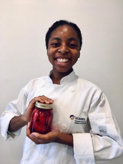 Angelique Ricketts is dressed in a chefs coat, holding a jar of red preserves