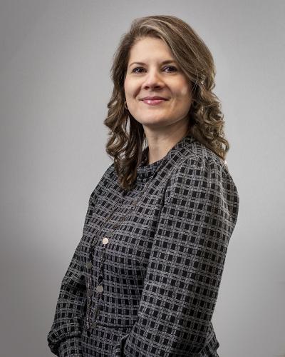 Irene Kosmas stands against a grey background, smiling over her shoulder, wearing a grey suit jacket