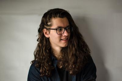 Stephen Bykowy is in front of a white background, with his head turned to the side. He is wearing glasses and a black shirt