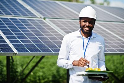 A man smiles in front of a giant solar panel with a white hard hat and white shirt, holding a clipboard