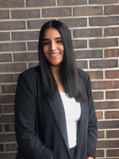 Nikita Ravi smiles, standing in front of a brick wall. She has long brown hair and is wearing a black blazer