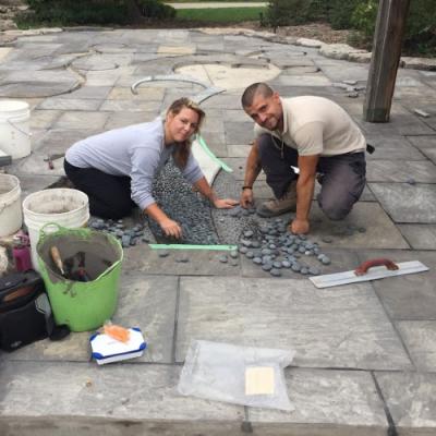 Two people are hunched over, working on a mosaic tile on the cobblestoned ground