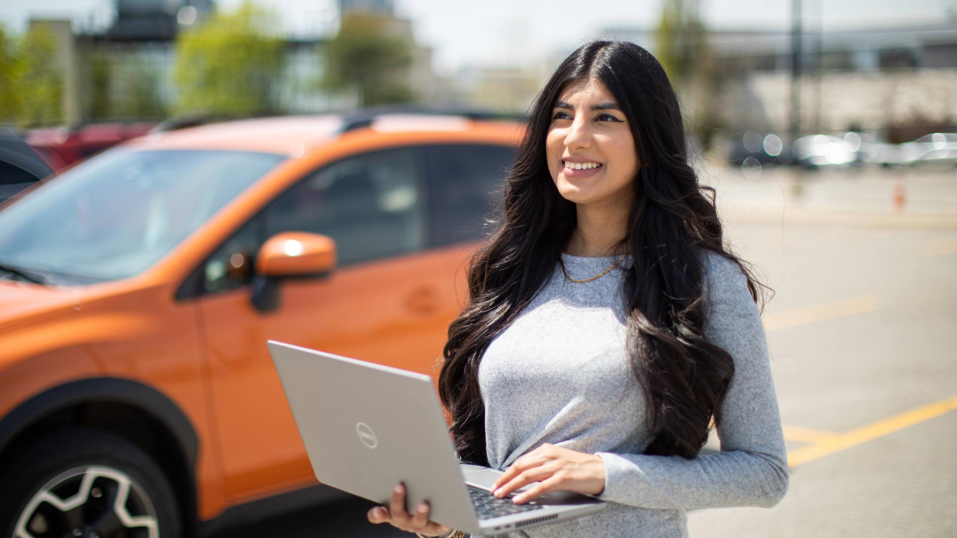 Kiranvir Kaur standing in front of an orange car, holding a laptop