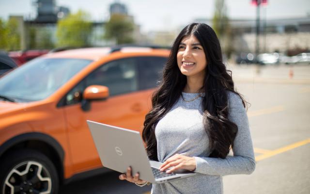 Kiranvir Kaur standing in front of an orange car, holding a laptop